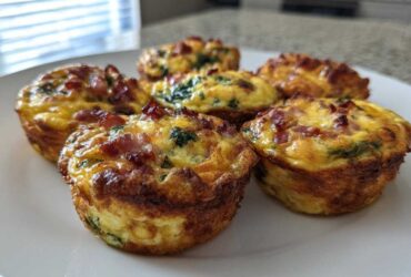 A close-up of six golden-brown Egg Muffin Cups topped with crispy bacon and visible spinach on a white plate.