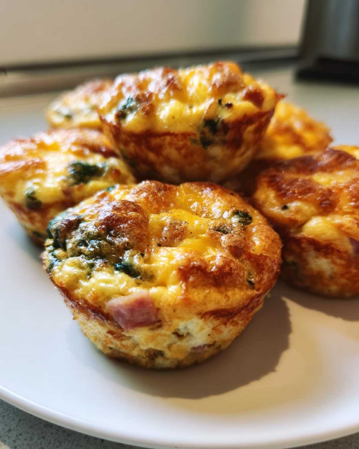 Close-up of several golden brown Egg Muffin Cups piled on a white plate, showing visible bits of ham and spinach.