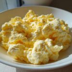 Close-up of incredibly fluffy scrambled eggs served in a white bowl, highlighted by bright sunlight.