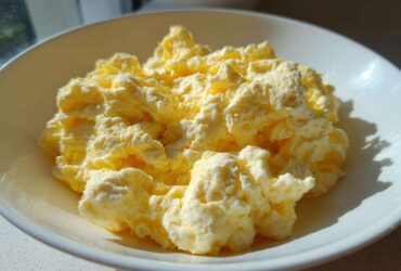 Close-up of incredibly fluffy scrambled eggs served in a white bowl, highlighted by bright sunlight.