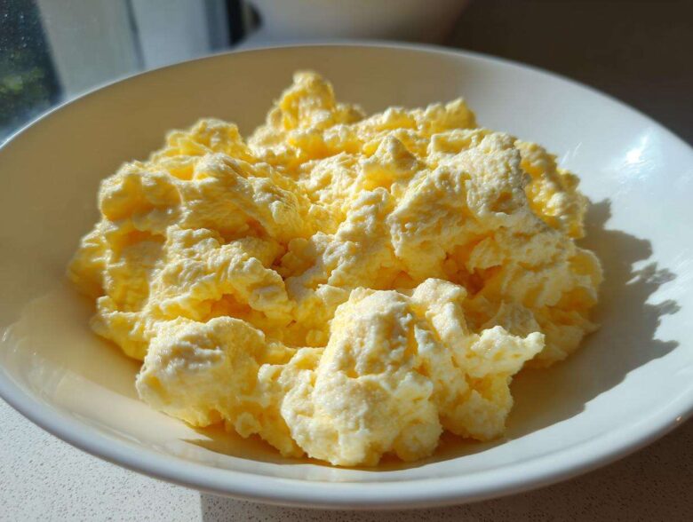 Close-up of incredibly fluffy scrambled eggs served in a white bowl, highlighted by bright sunlight.