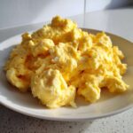 Close-up of perfectly fluffy scrambled eggs piled high on a white plate, showcasing a creamy texture.