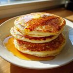 A stack of three fluffy sourdough pancakes topped with melting butter and dripping with syrup.