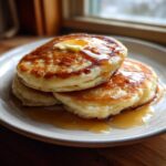 A stack of three fluffy sourdough pancakes topped with melting butter and drizzled with maple syrup.
