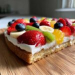 Close-up of a slice of fruit pizza sugar cookie topped with cream cheese frosting and colorful fresh fruit.