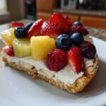 A slice of fruit pizza sugar cookie topped with cream cheese frosting, strawberries, blueberries, raspberries, and pineapple chunks.