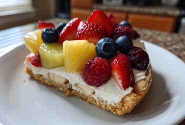 A slice of fruit pizza sugar cookie topped with cream cheese frosting, strawberries, blueberries, raspberries, and pineapple chunks.