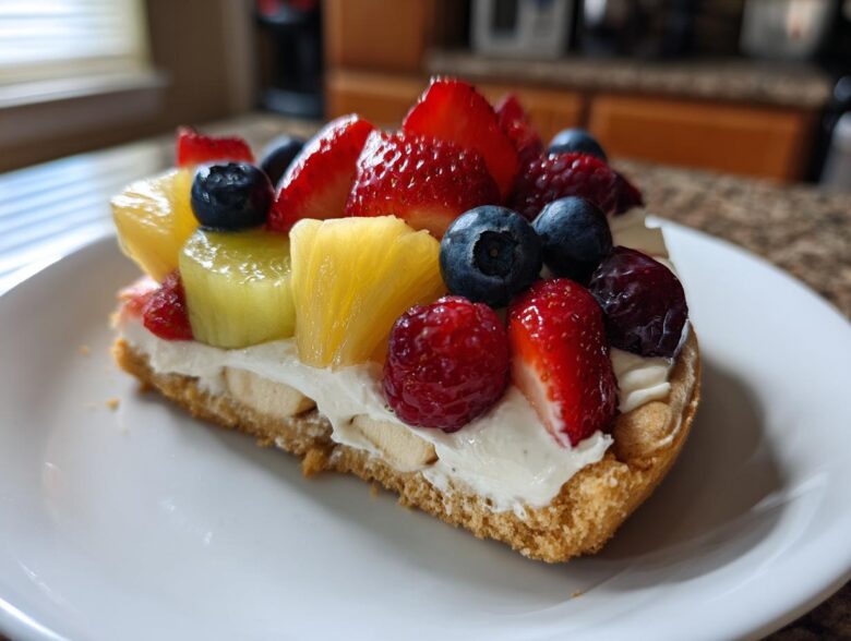 A slice of fruit pizza sugar cookie topped with cream cheese frosting, strawberries, blueberries, raspberries, and pineapple chunks.