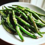 A close-up of vibrant, glossy green beans tossed with minced garlic served on a white plate.