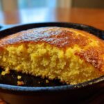 Close-up of golden, moist skillet cornbread baked in a cast iron pan with a piece removed.
