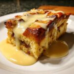 A decadent slice of bread pudding topped with rich vanilla sauce, served on a white plate.