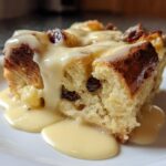 Close-up of a square slice of rich bread pudding studded with raisins, topped generously with vanilla cream sauce.