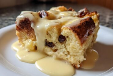 Close-up of a square slice of rich bread pudding studded with raisins, topped generously with vanilla cream sauce.