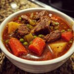 Close-up of a white bowl filled with rich, savory vegetable beef soup featuring large chunks of beef, carrots, potatoes, and peas.