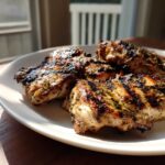 Four pieces of perfectly grilled greek chicken with visible char marks and herbs served on a white plate.