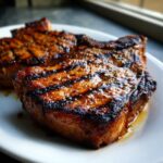 Close-up of two juicy, grilled pork chops showing dark char marks from the grill, coated in a savory marinade for pork chops.