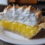 A close-up slice of lemon meringue pie showing thick yellow filling and tall, toasted meringue.