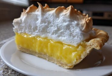 A close-up slice of lemon meringue pie showing thick yellow filling and tall, toasted meringue.