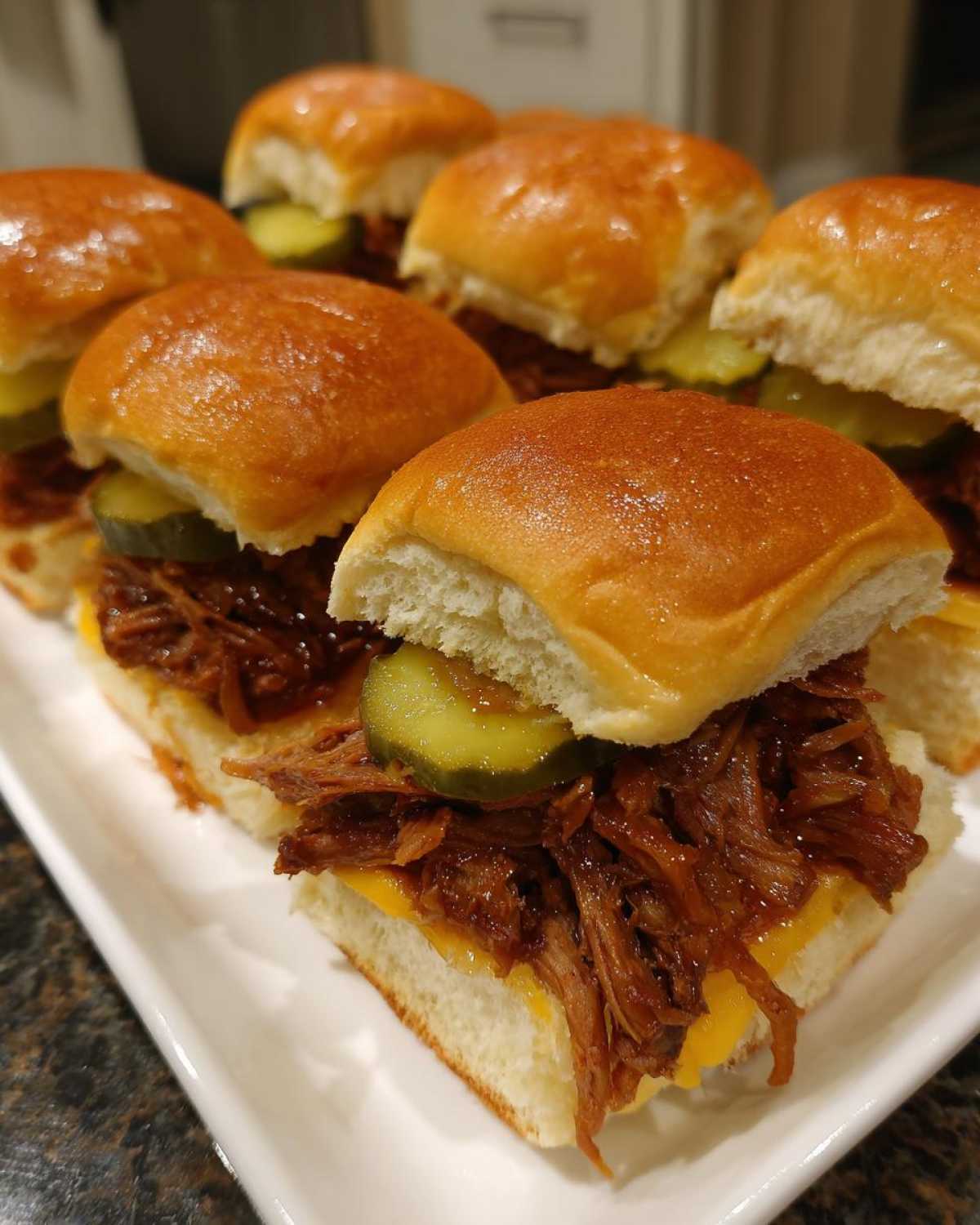 Close-up of several delicious mississippi pot roast sliders topped with melted cheese and pickles on shiny buns.