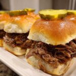 Close-up of several delicious mississippi pot roast sliders piled high with shredded beef and topped with a pickle slice.