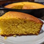 A close-up of a moist slice of buttermilk cornbread on a white plate, with the rest of the cornbread in a cast iron skillet blurred in the background.
