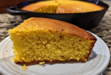 A close-up of a moist slice of buttermilk cornbread on a white plate, with the rest of the cornbread in a cast iron skillet blurred in the background.