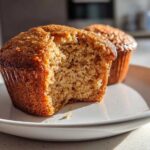 Close-up of a moist honey bran muffin with a bite taken out, showing the crumb texture, on a white plate.