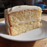 A close-up of a moist, two-layer slice of vanilla cake recipe covered in thick white frosting, sitting on a white plate.
