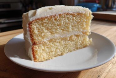A close-up of a moist, two-layer slice of vanilla cake recipe covered in thick white frosting, sitting on a white plate.