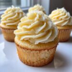 Close-up of a moist vanilla cupcake topped with a swirl of creamy vanilla frosting, with more vanilla cupcakes blurred in the background.