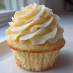 Close-up of a single, moist vanilla cupcake topped with a tall swirl of creamy vanilla buttercream frosting.
