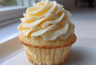 Close-up of a single, moist vanilla cupcake topped with a tall swirl of creamy vanilla buttercream frosting.