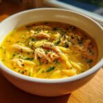 Close-up of a white bowl filled with bright yellow penicillin soup, featuring shredded chicken and orzo pasta, garnished with parsley.