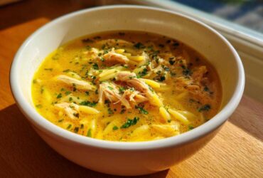 Close-up of a white bowl filled with bright yellow penicillin soup, featuring shredded chicken and orzo pasta, garnished with parsley.
