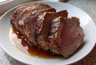Several thick slices of a perfectly cooked pork loin roast resting in its juices on a white plate.