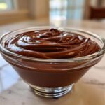 A close-up of rich, glossy homemade chocolate pudding swirled in a clear glass serving bowl.