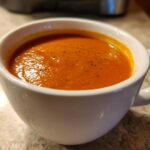 Close-up of a creamy, orange-red tomato bisque sprinkled with pepper in a white mug.
