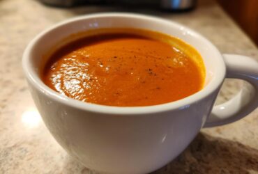 Close-up of a creamy, orange-red tomato bisque sprinkled with pepper in a white mug.