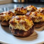 Five baked sausage stuffed mushrooms with a golden, cheesy, browned topping served on a white plate.