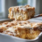 A close-up shot of a piece of fluffy Cinnamon roll casserole being lifted out of the baking dish, dripping with white icing.