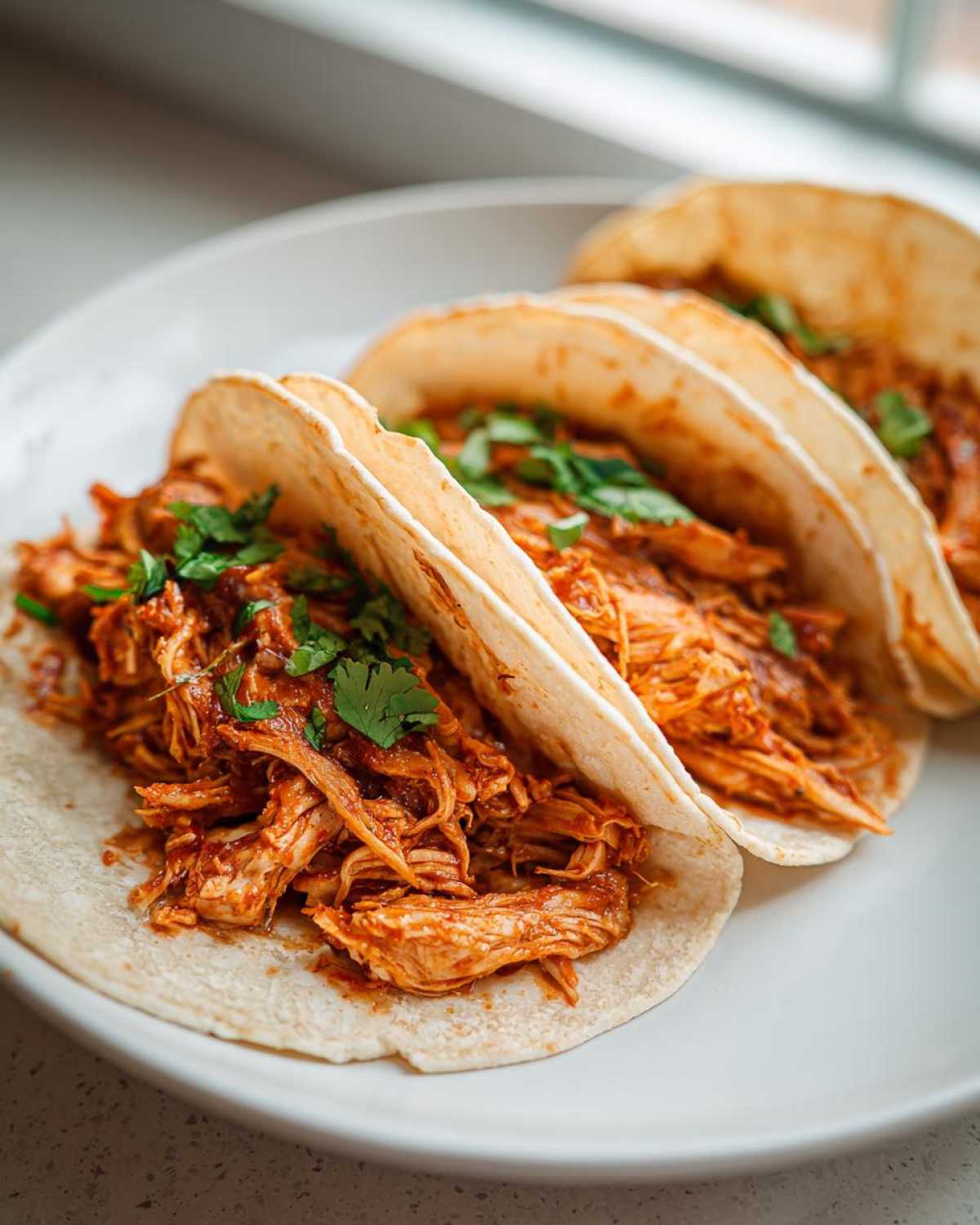 Three soft tortilla chicken tacos overflowing with saucy shredded chicken and topped with fresh cilantro.