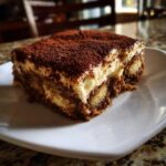 A close-up of a perfect slice of tiramisu recipe, showing layers of cream and coffee-soaked ladyfingers, dusted heavily with cocoa.