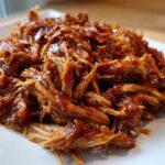 Close-up of saucy, shredded Slow cooker pulled pork piled high on a square white plate.