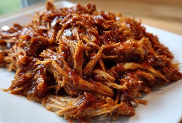 Close-up of saucy, shredded Slow cooker pulled pork piled high on a square white plate.