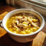 A steaming white bowl filled with golden penicillin soup, featuring shredded chicken and small ring pasta.
