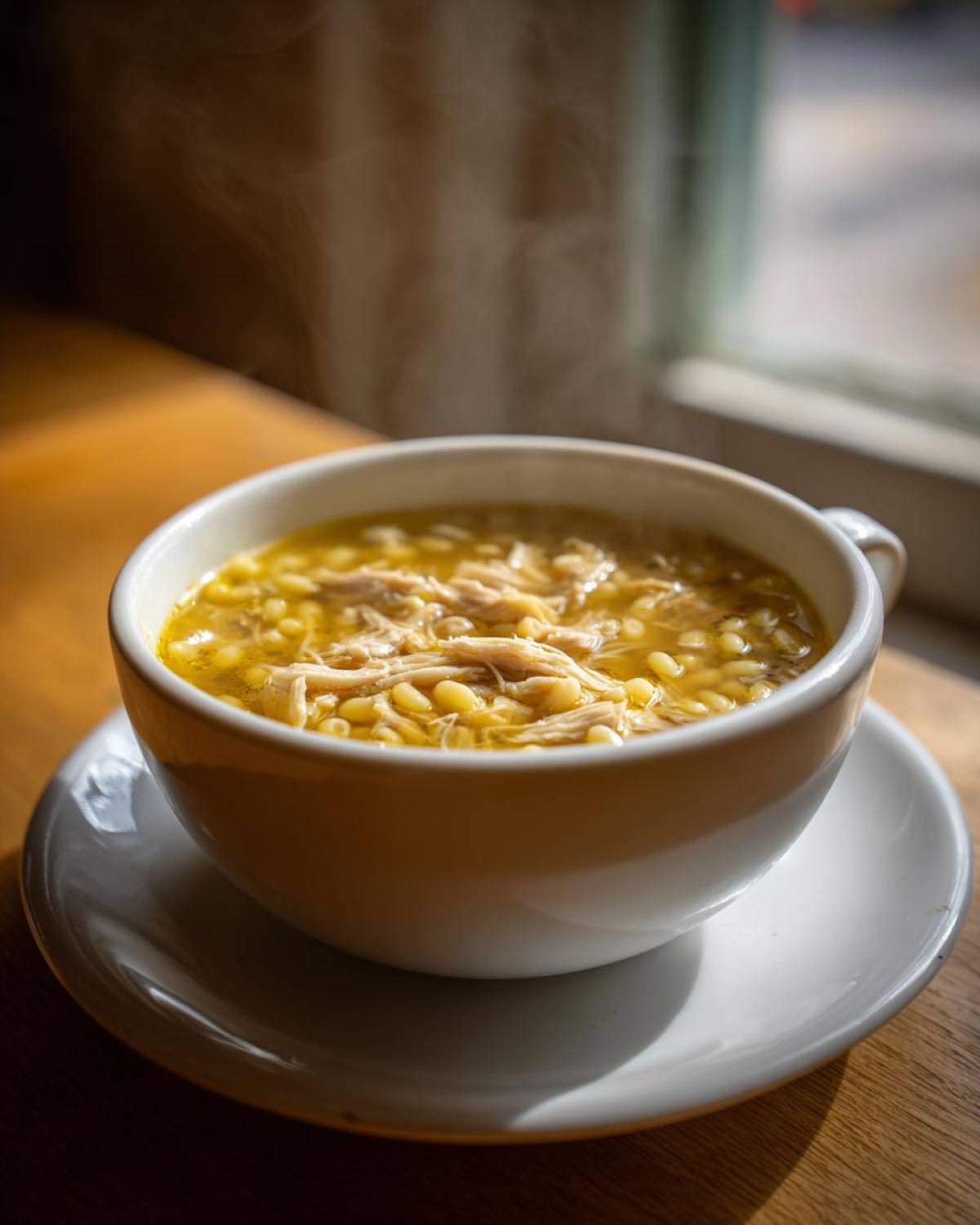 A steaming white bowl of penicillin soup filled with shredded chicken and small pasta sits on a saucer by a sunlit window.