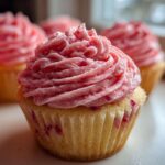 A close-up of a freshly baked strawberry cupcake topped with vibrant pink strawberry frosting, with more strawberry cupcakes blurred in the background.