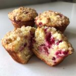 One of the strawberry muffins is cut in half, showing the moist interior filled with bright red fruit and topped with golden streusel.
