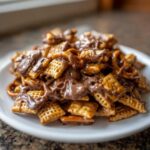 Close-up of toffee chex mix recipe piled on a white plate, covered in melted chocolate drizzle.