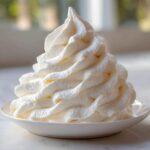 Close-up of a tall, perfectly piped swirl of creamy white vanilla buttercream frosting on a small white plate.